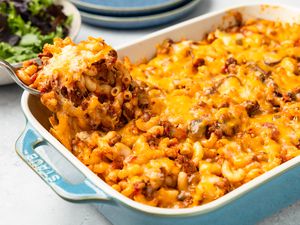 Spoonful of Johnny Marzetti Casserole lifted from the casserole dish, and in the surroundings, a stack of plates and a bowl of mixed greens salad