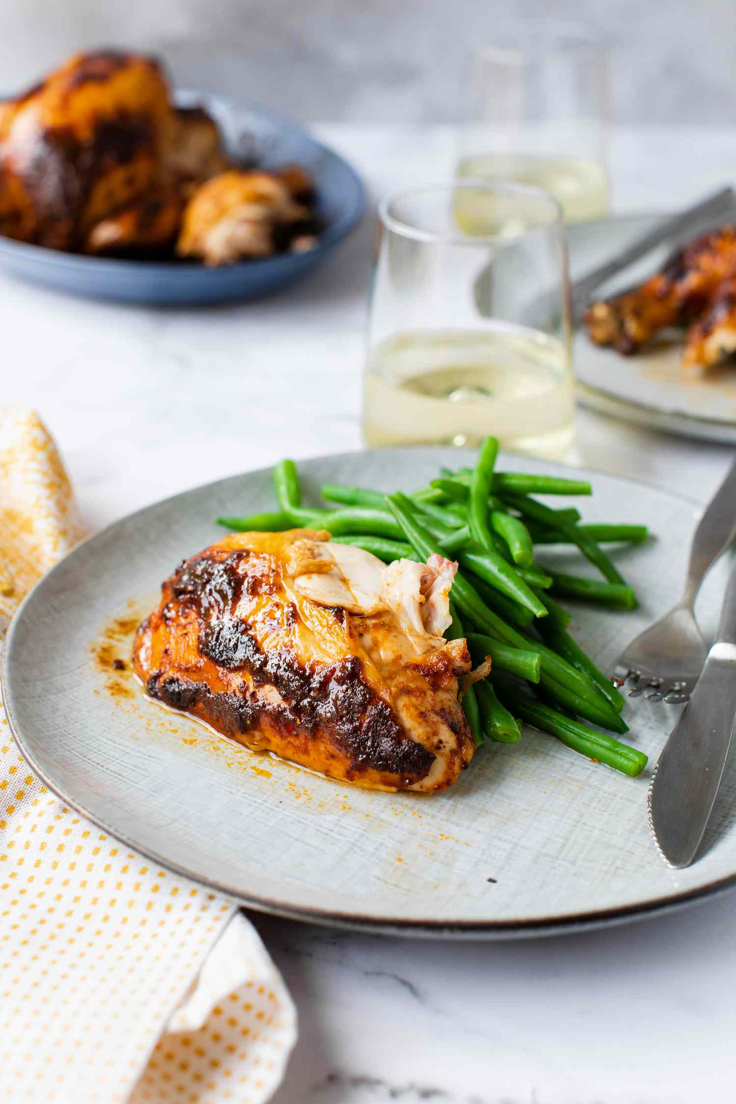 Plate of Smoked Paprika Roasted Chicken with Green Beans, Surrounded by a Table Setting with Glasses, Table Napkins, and More Plates of Chicken