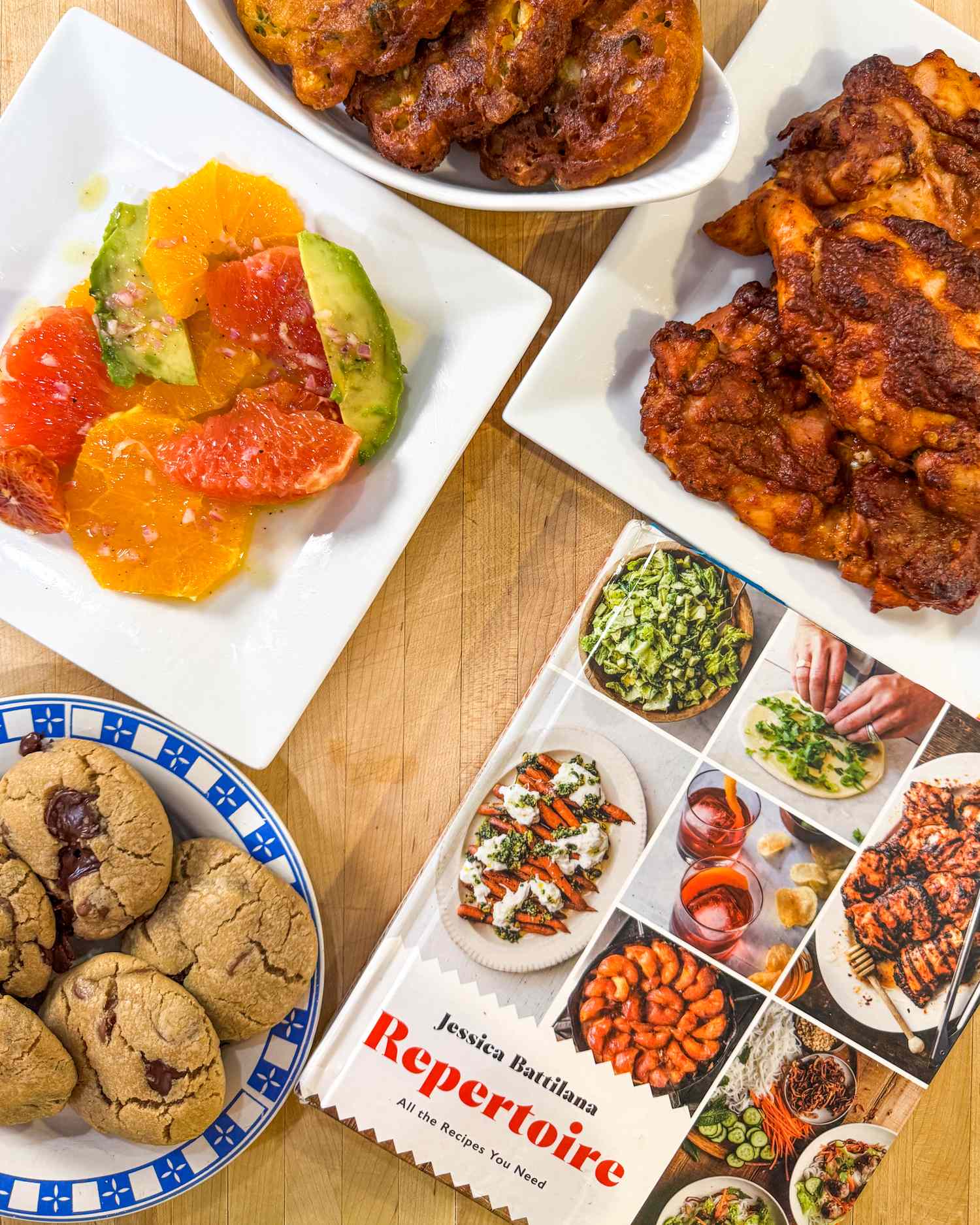 A cookbook titled Repertoire surrounded by various plated dishes including cookies fruit salad and fried chicken