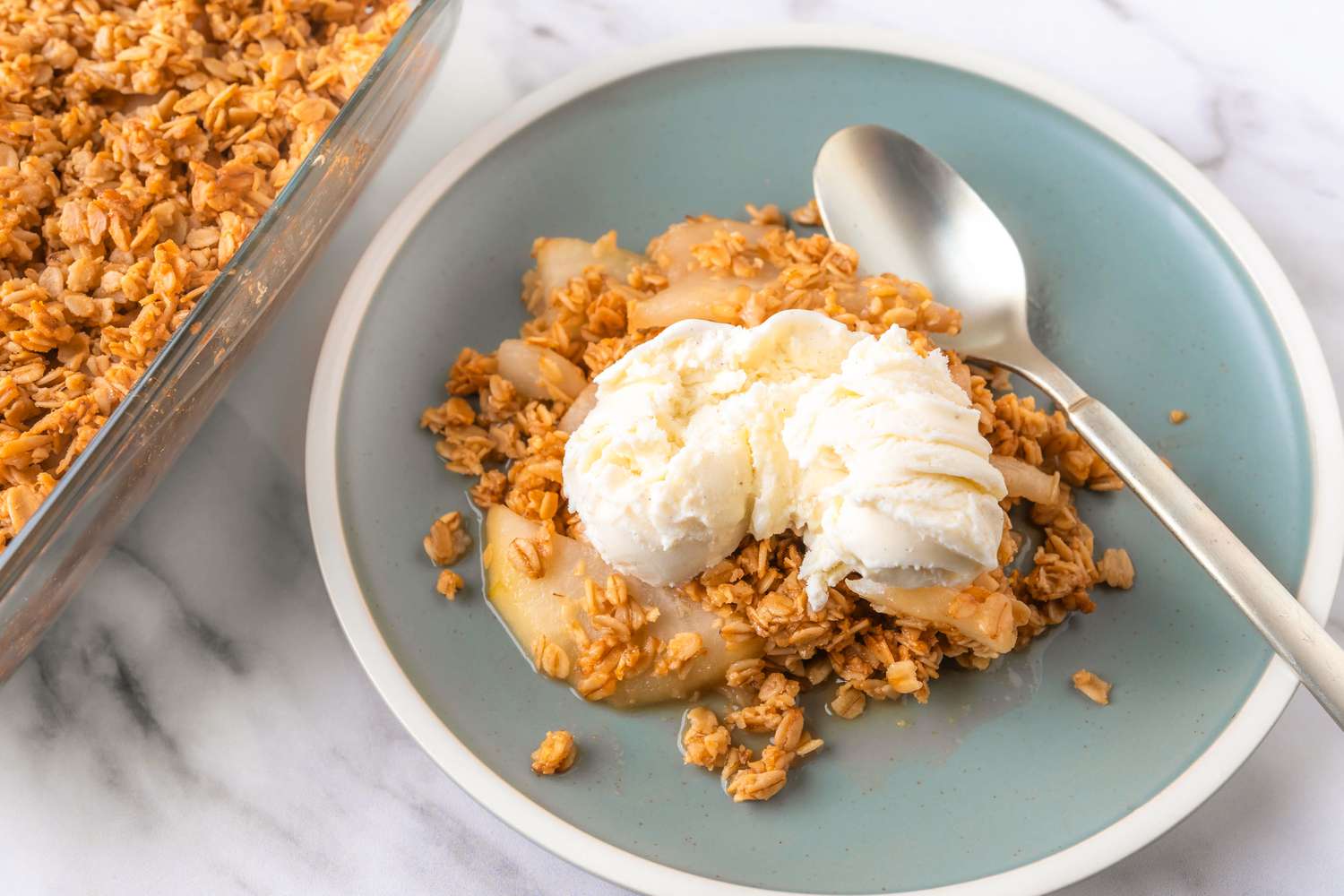 Overhead view of a teal plate of 5-ingredient vanilla pear crisp recipe topped with vanilla ice cream and next to a spoon and the baking dish from the crisp