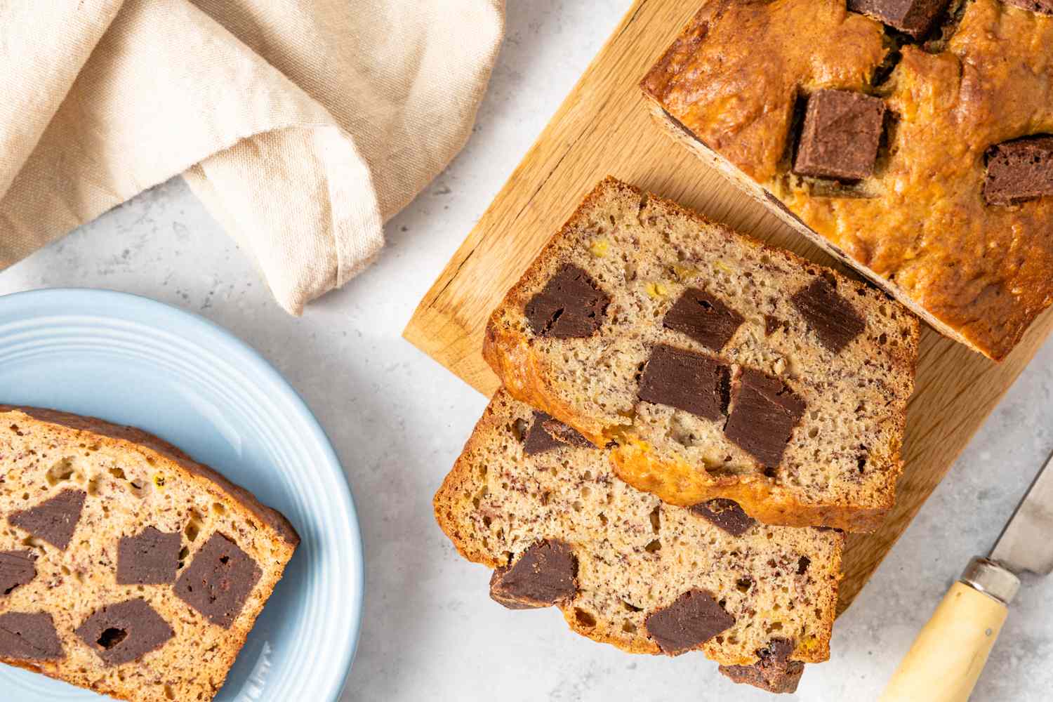 On a Wooden Board, Loaf of Brownie Banana Bread with Slices Cut Off, and Next to It, a Plate with a Slice, a Kitchen Towel, and a Knife