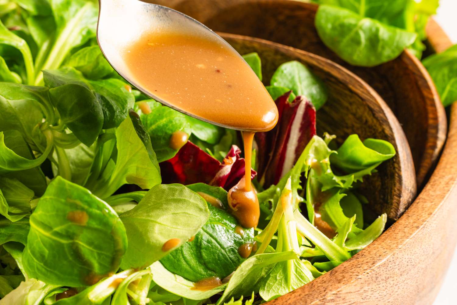A spoon drizzling a dressing onto a bowl of mixed greens salad