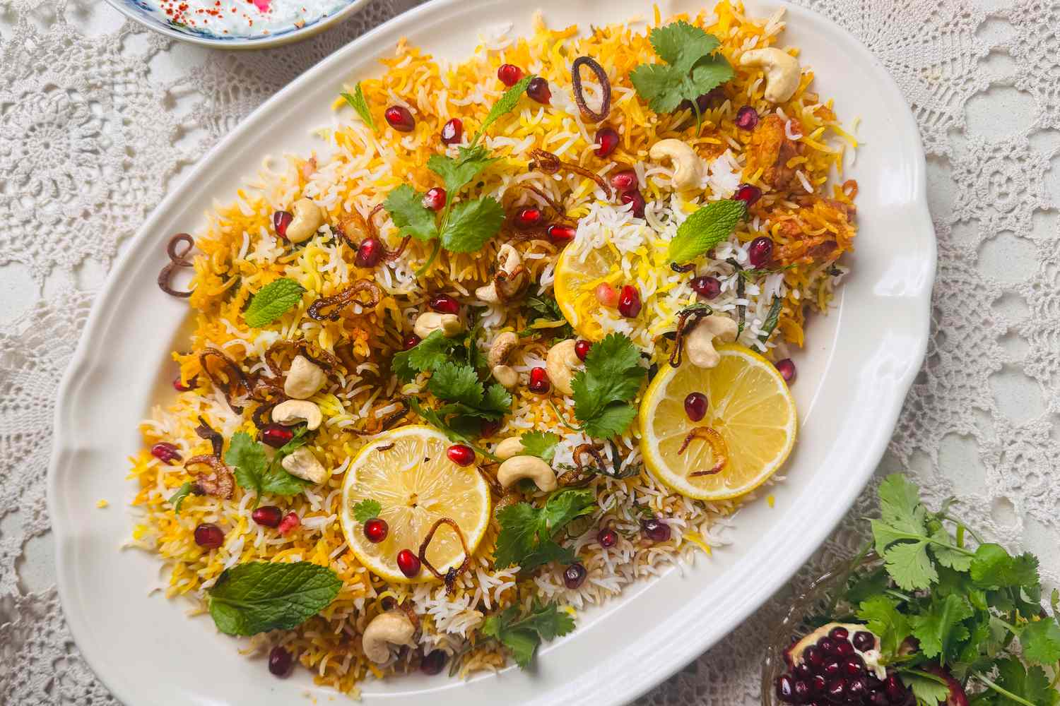 Chicken biryani on a large, white porcelain serving platter. The rice dish is garnished with lemon slices, pomegranate arils, cashews, and mint and cilantro leaves.