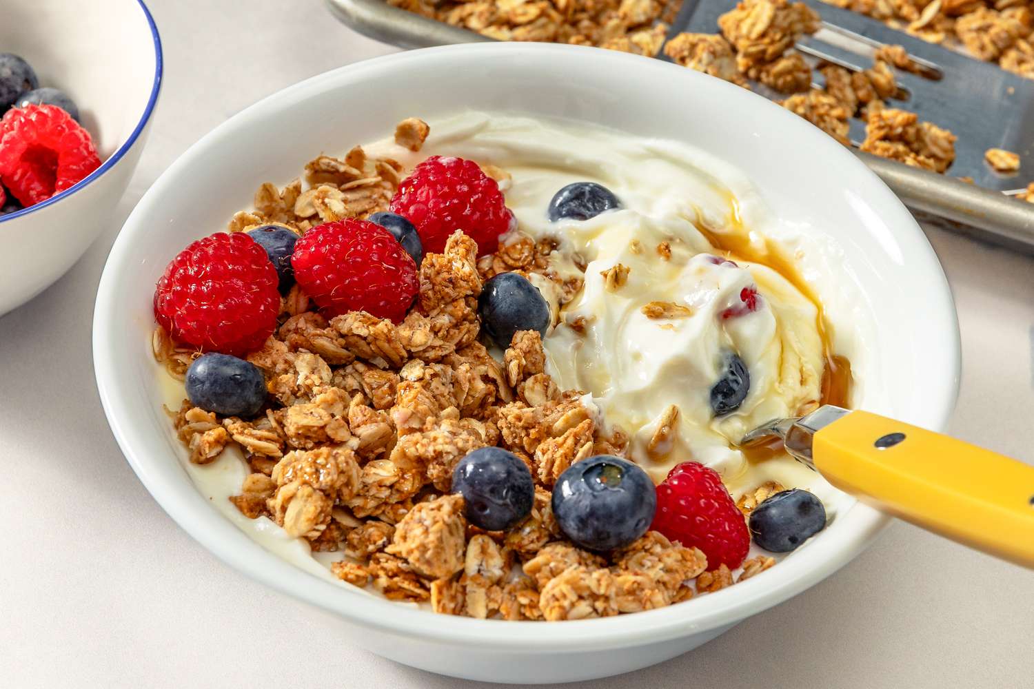 Angled view of a bowl of granola topped with blueberries, strawberries, yogurt and maple syrup with a yellow handled spoon on a white surface