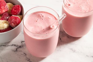 Two glasses of pink smoothie with a glass straw next to a bowl of frozen strawberries and banana slices