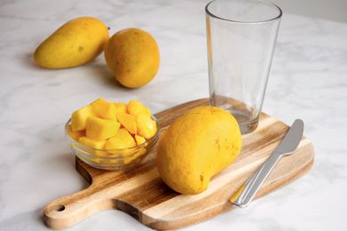 Sliced mango pieces in a bowl on a cutting board with a whole mango a glass and a knife nearby