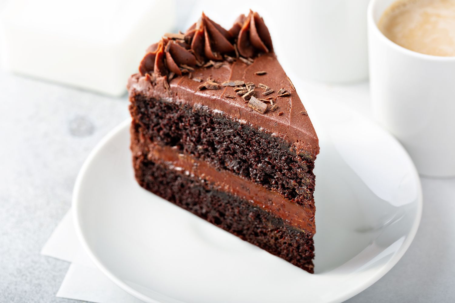 A slice of chocolate layer cake on a white plate topped with frosting and chocolate shavings next to a cup of coffee