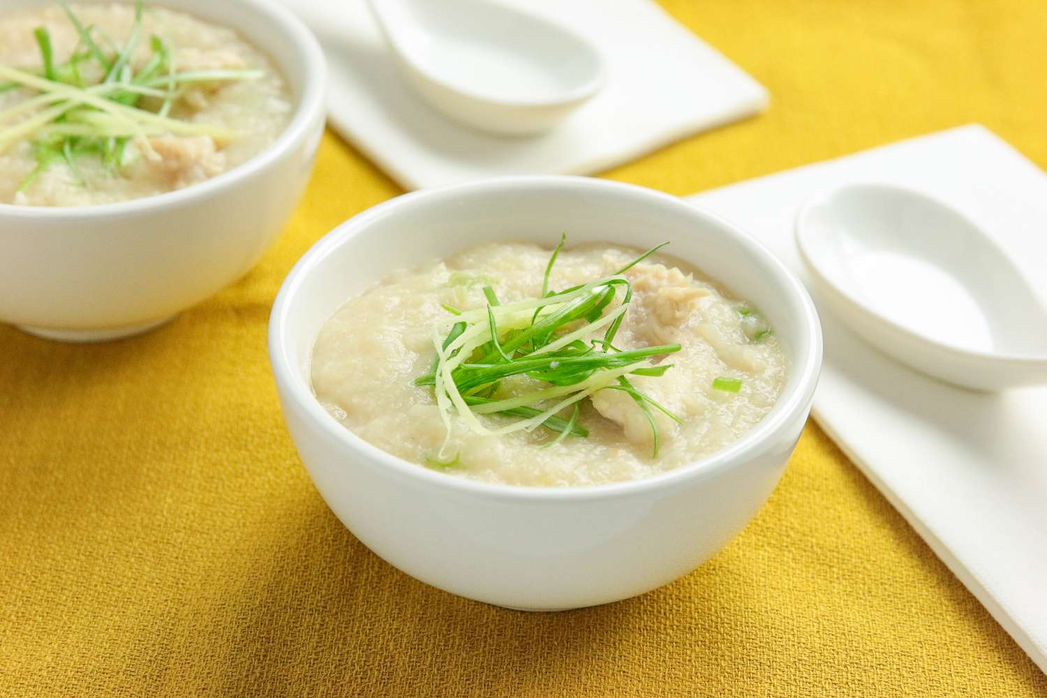 Two Bowls of Chicken Congee Topped with Sliced Ginger and Sliced Green Onion, and Next to the Bowls, Two Soup Spoons on Table Napkins, All on a Mustard Colored Kitchen Towel