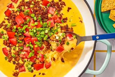 Queso dip with toppings of chopped tomatoes green onions and chorizo served with a spoon and tortilla chips on the side