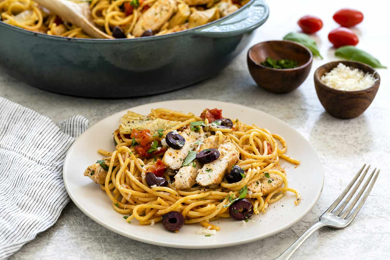 Side view of a plate of one pot chicken pasta dinner. A large skillet is behind the plate of pasta and grape tomatoes, basil and seasoning are in the upper right.