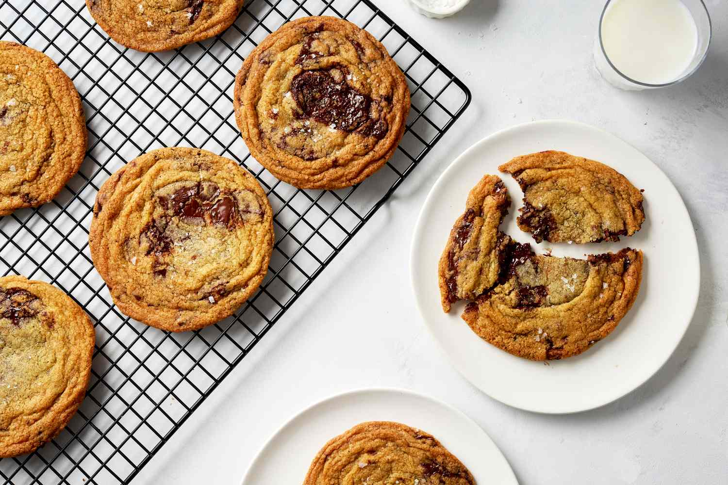 Chocolate chip cookies on a cooling rack and plates with a glass of milk nearby
