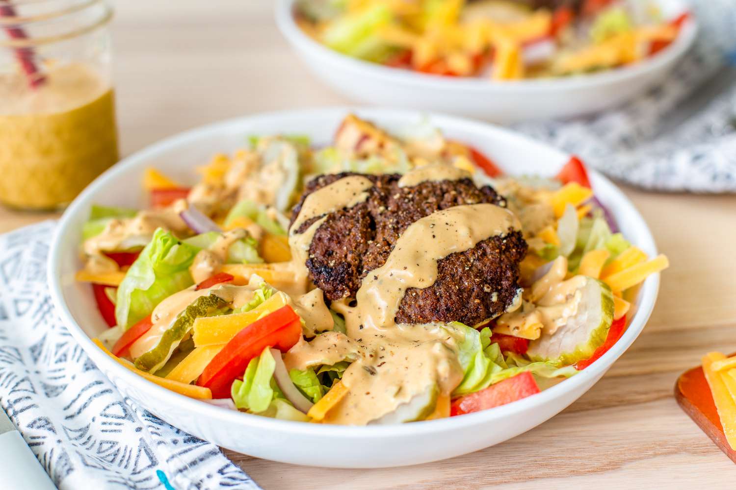 Bowl of Burger Salad at a Table Setting With Another Bowl of Salad in the Background, a Mason Jar of Sauce, and Table Napkins