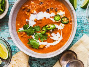 Creamy Chicken Enchilada Soup Topped with Jalapenos, Cilantro, and Crema in a Bowl at a Table Setting with Two Spoons on a Table Napkin, Bowls of Toppings (Jalapenos and Lime Wedges), and Tortillas