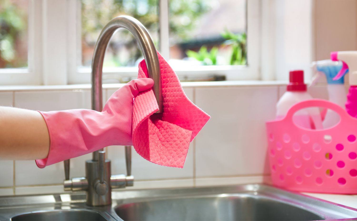 An arm reaching into frame with a pink cleaning cloth. The hand is wearing a pink dish glove and is using the cleaning cloth to polish a kitchen faucet.