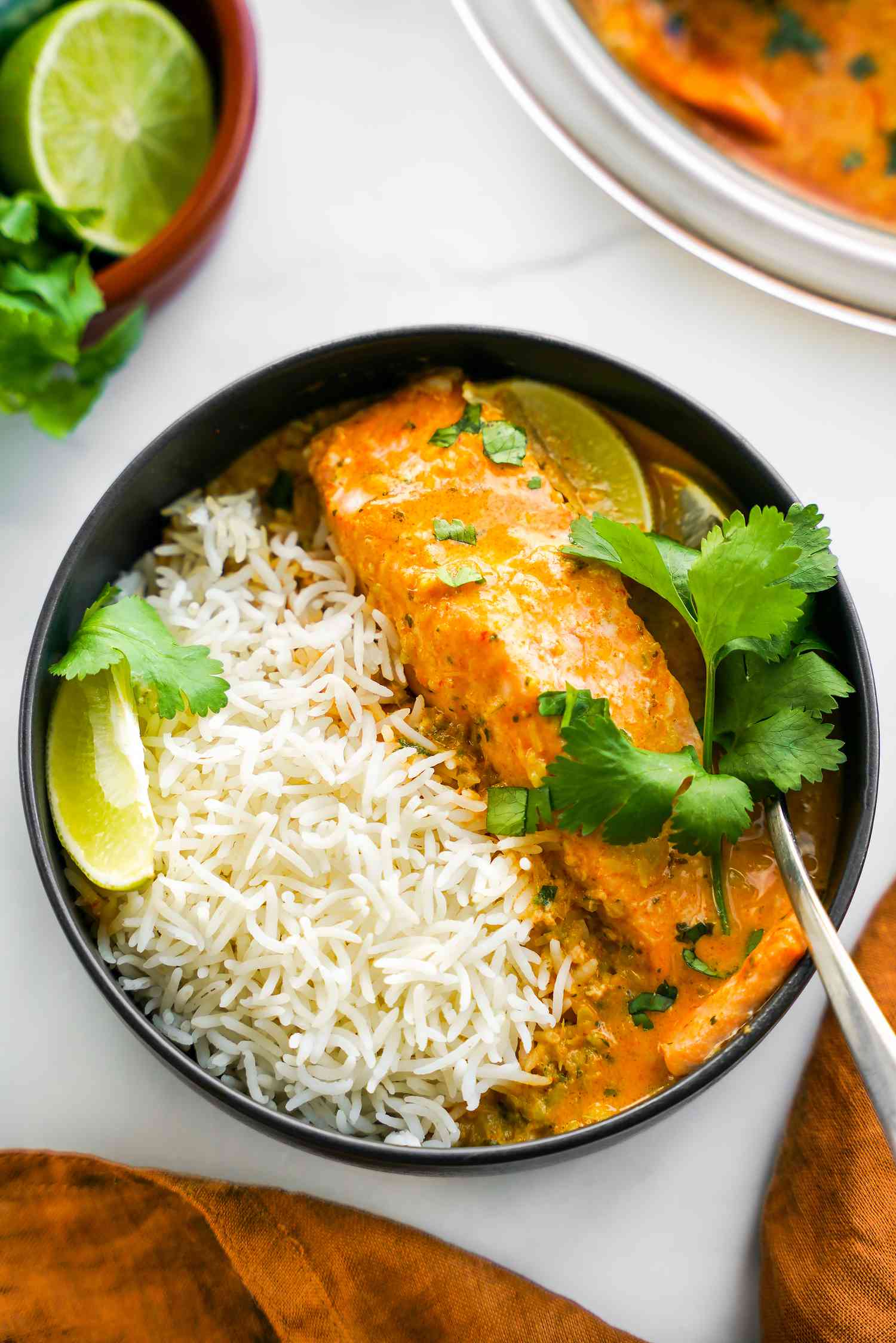 Bowl of Microwave Red Curry Poached Salmon with Rice and Garnished with Cilantro and Lime Wedge, and in the Surroundings, a Kitchen Towel, a Bowl with More Curry, and a Bowl with a Halved Lime and Cilantro