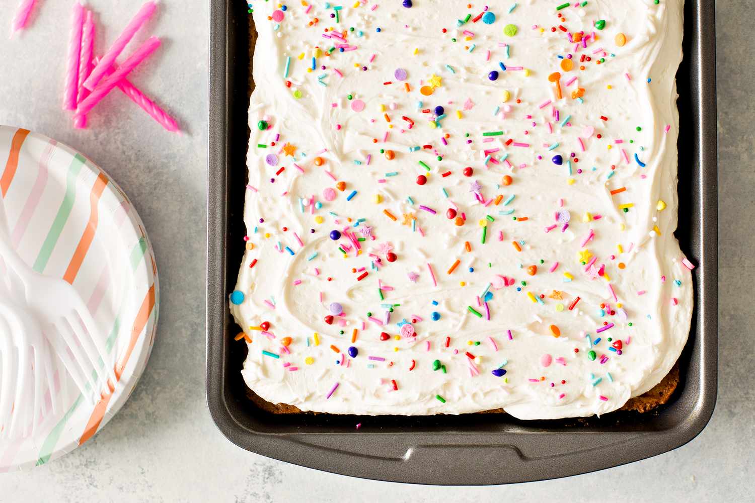 A frosted sheet cake with sprinkles in a rectangular pan forks and decorative candles on the side
