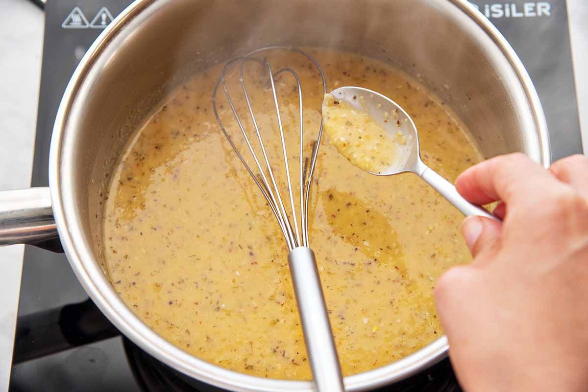 Creamy golden sauce in a pan with a whisk. Hand holding a spoon dripping sauce over pan.