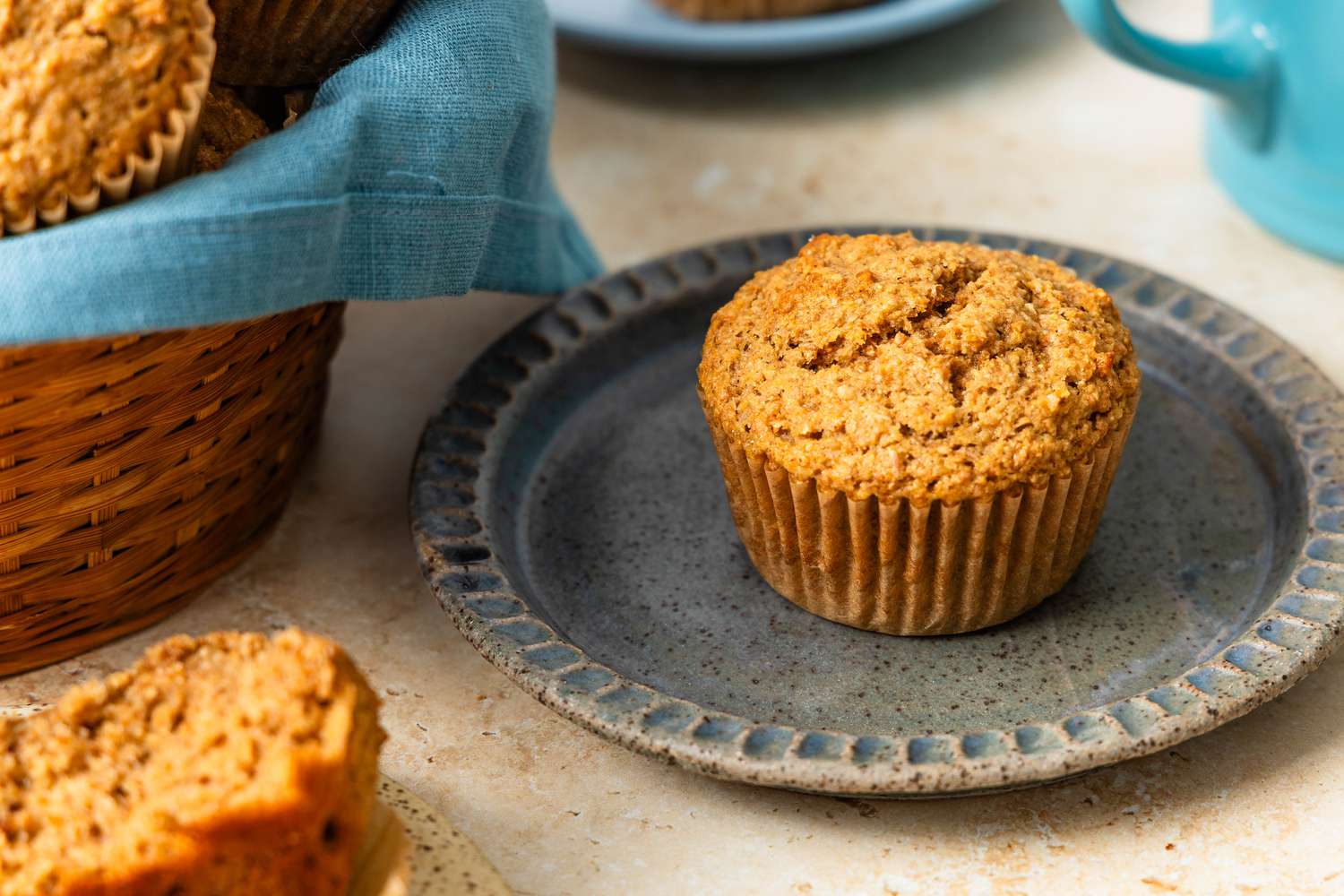 Bran muffin on a plate, and in the surrondings, another muffin on a plate, a table napkin lined basket, and a kettle