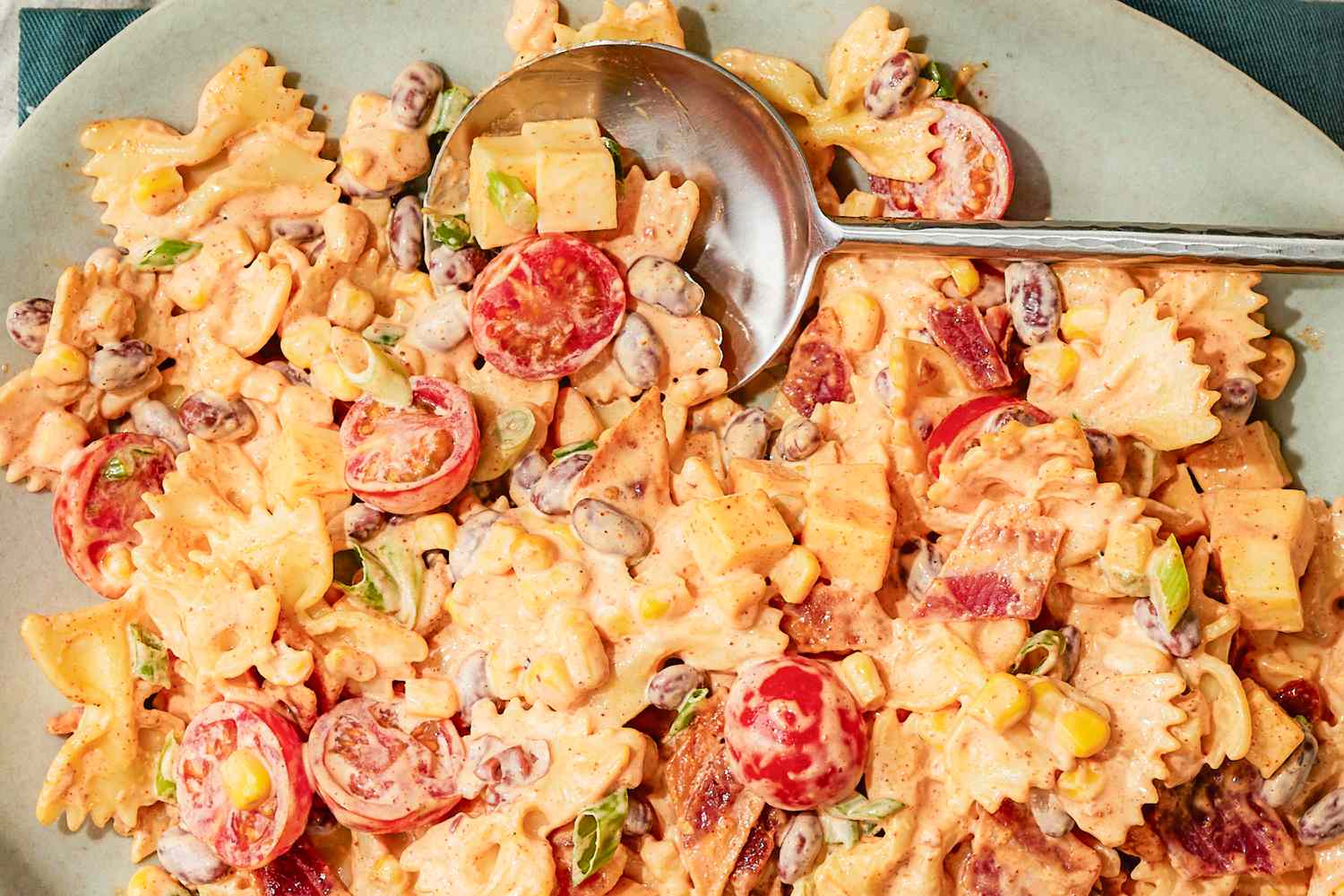 A serving of pasta salad with a variety of ingredients including cherry tomatoes, beans, and bacon, served with a spoon on the side