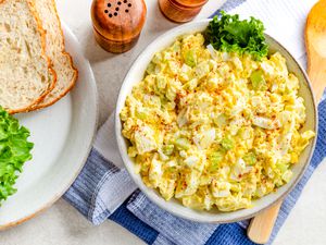 bowl of egg salad with a piece of lettuce on the counter next to a plate with bread and lettuce, salt and pepper shakers, and large spoon on a table napkin