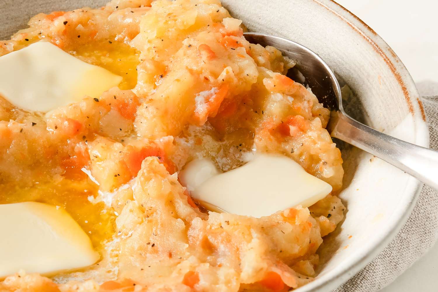 A dish of mashed potatoes and carrots served with pats of melting butter placed on a white bowl with a spoon