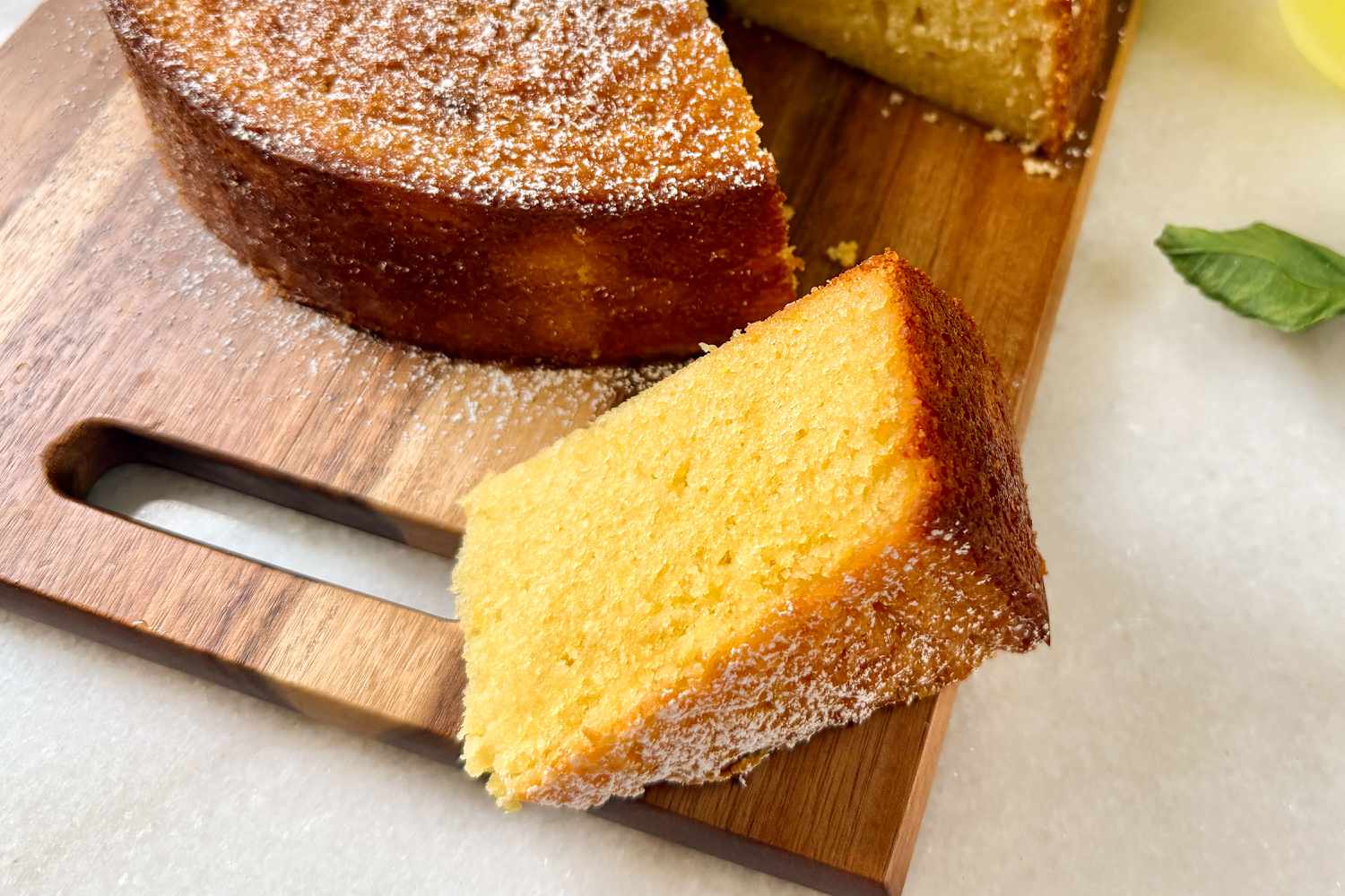Meghan Markle's Lemon Olive Oil Cake slice on a cutting board with the rest of the cake in the background