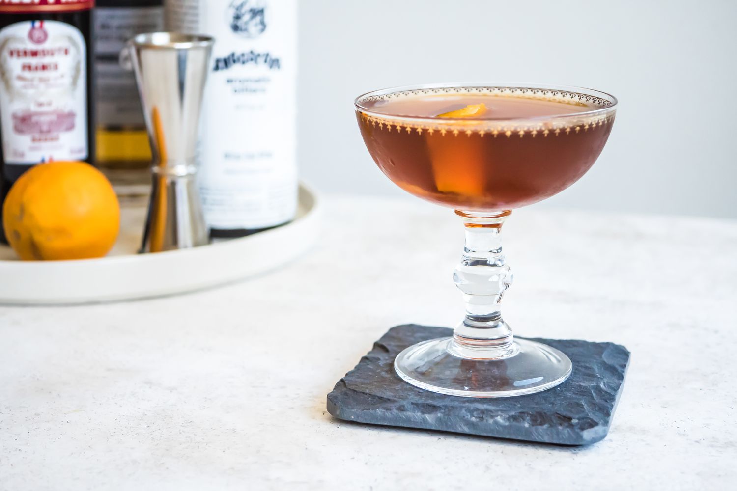 Rob Roy in Cocktail Glass on Top of Granite Coaster Surrounded by a with a Tray with Recipe Ingredients (Vermouth, Whiskey, and Orange) in the Background
