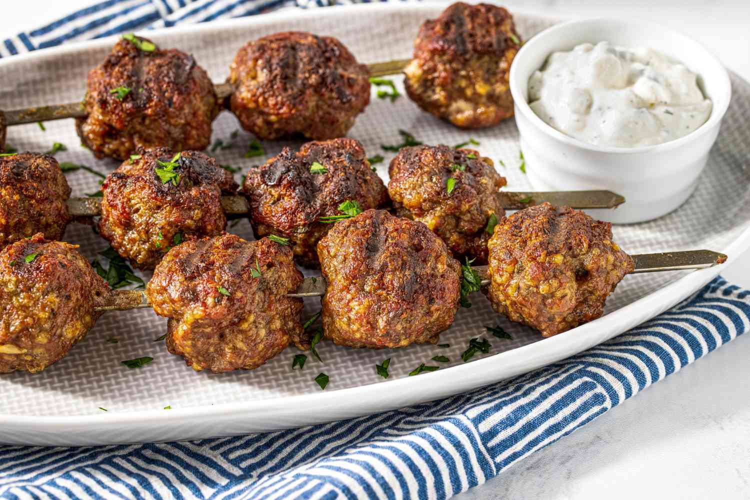 Platter of Grilled Meatballs With a Small Bowl of Tzatziki, All on a Blue and White Stripped Kitchen Towel