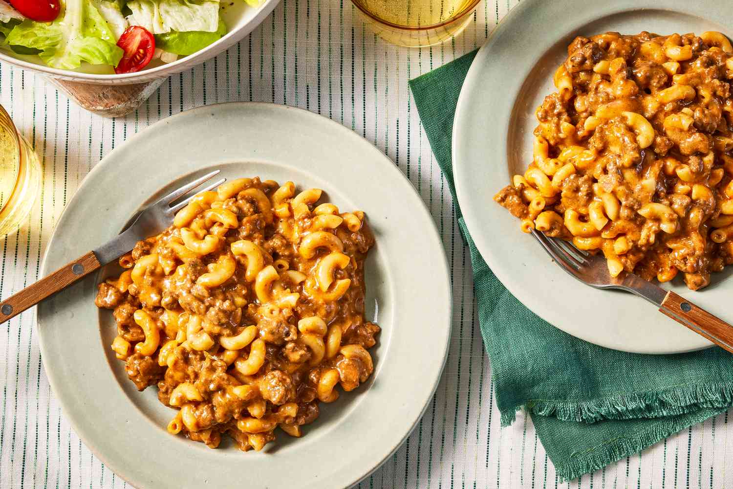 homemade hamburger helper on two plates at a table setting with two glasses of soda and a side salad