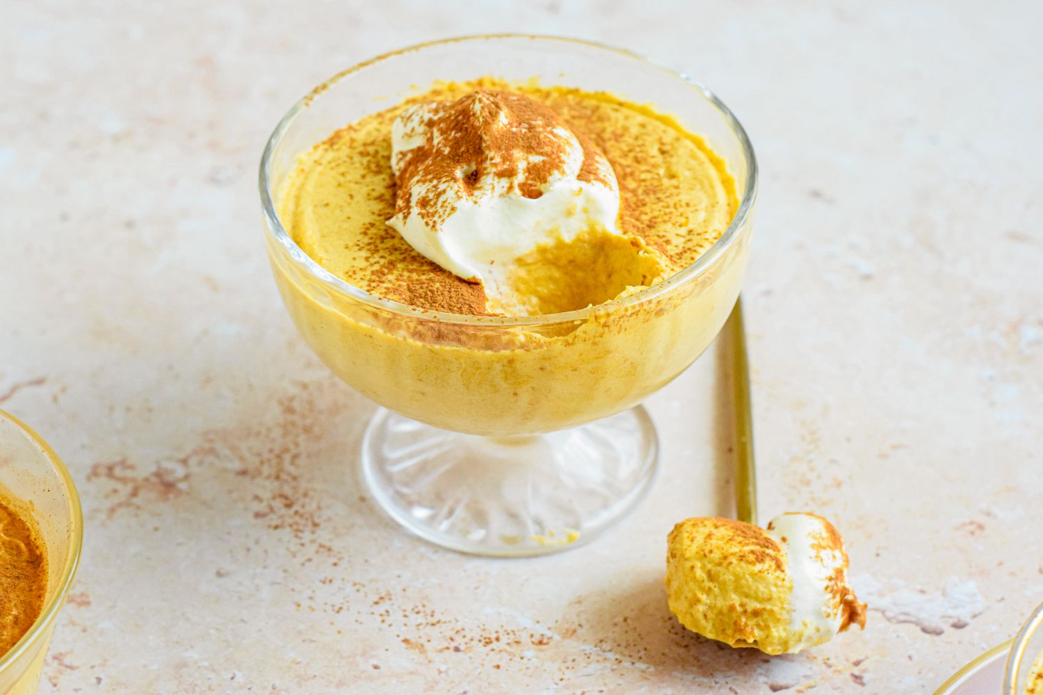 2-ingredient pumpkin mousse topped with whipped cream and a sprinkle of pumpkin spice, all in a sherbert glass. On the counter, a spoonful of mousse.