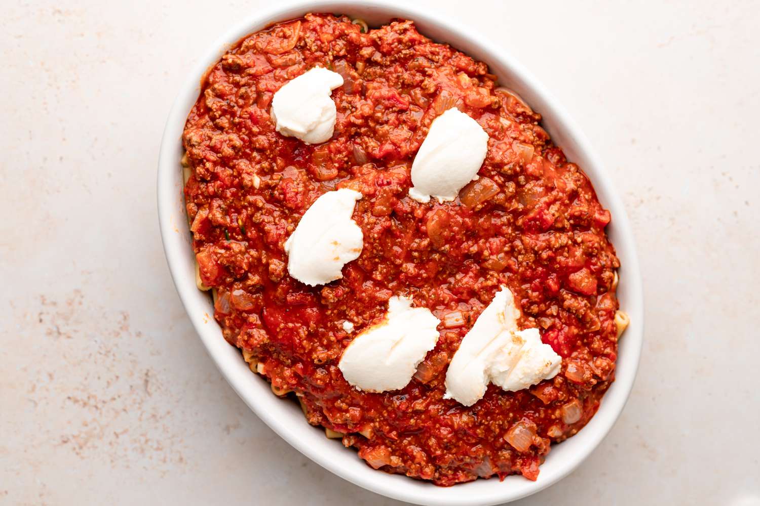 Ziti noodles in an oval baking dish and topped with a meat sauce and dollops of cheese.