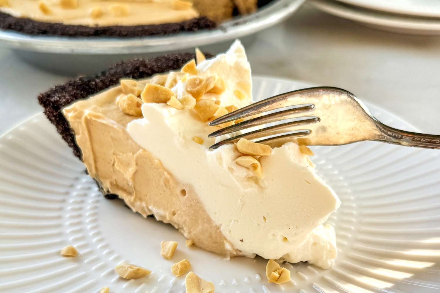 A slice of peanut butter pie with whipped topping and crushed peanuts on a plate a fork resting on the slice surface