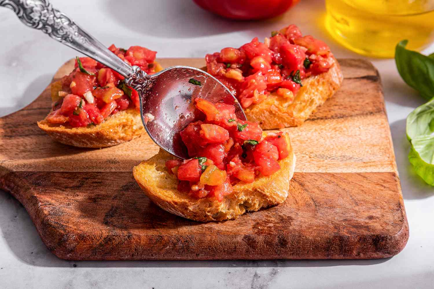 Bruschetta topping added to toasted baguette slice using a spoon. Baguette slice on a small wooden board with more bruschetta with tomato and basil, and in the surroundings, a bottle of olive oil, a tomato, and a piece of basil