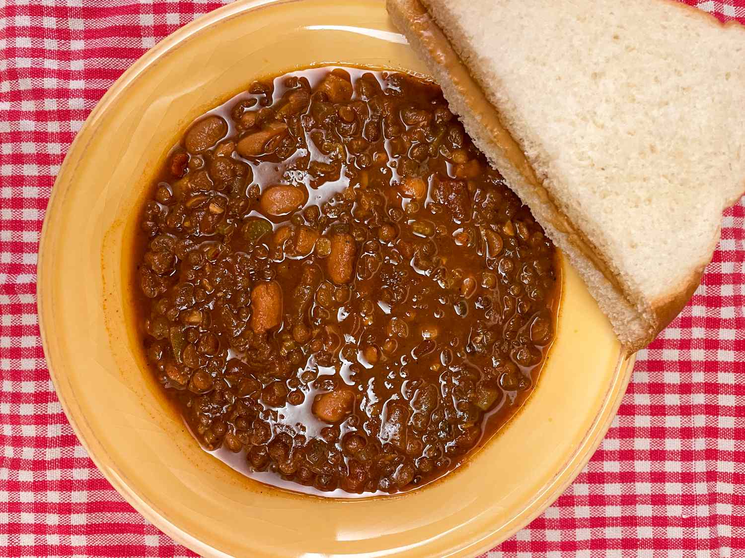 A bowl of chili with beans next to a peanut butter sandwich on a checkered surface