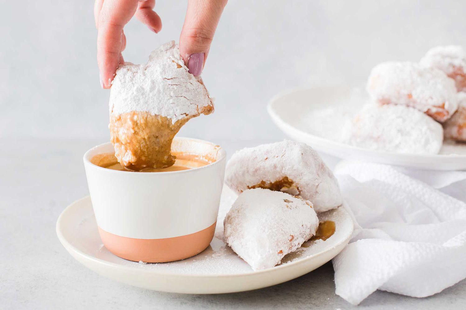 Powdered sugar donuts are on a cream plate and one is dipped in coffee. A platter of more donuts is behind and to the right.