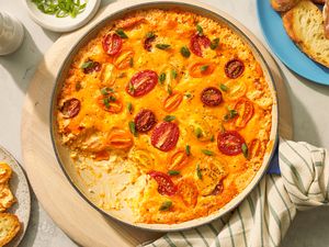 Tomato pie dip in a pan at a table setting with servings of dip on two plates with toasted baguette pieces, a bowl with sliced scallions, and a pepper grinder 