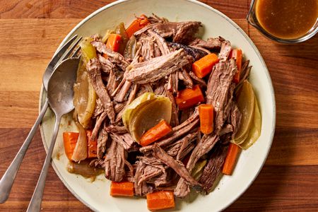 Overhead view of a plate of pot roast with carrots and onions and a fork and spoon all next to a cup of gravy on a wooden tabletop