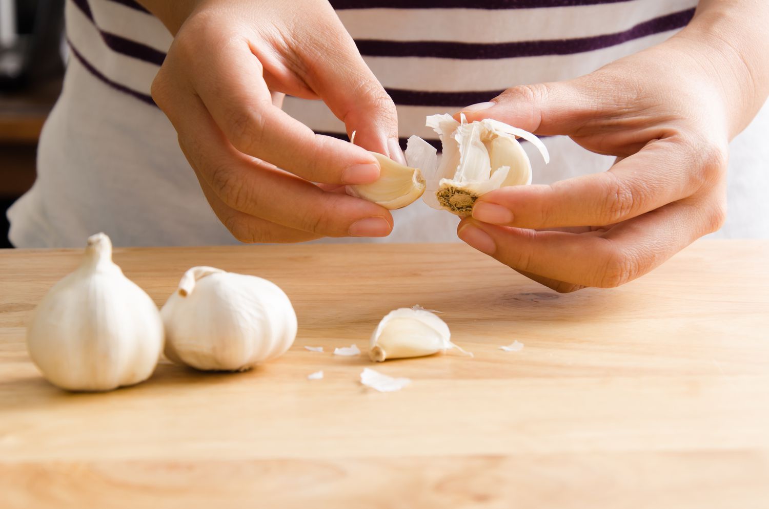 Person peeling a garlic, with more garlic cloves on a wooden counter below their hands