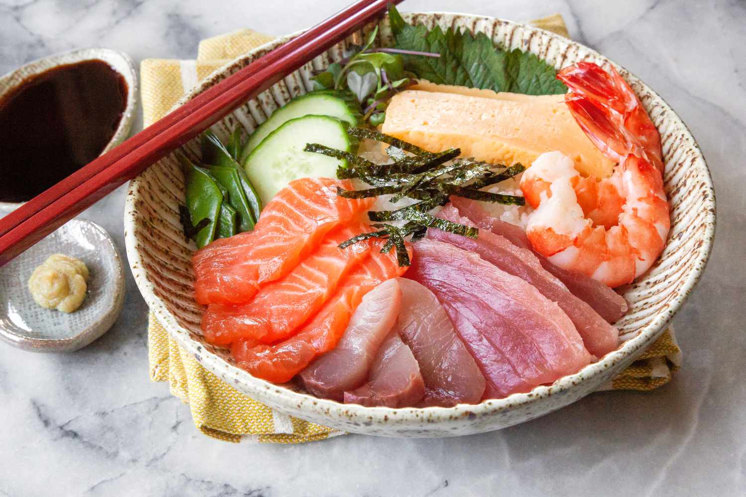 Chirashi Bowl with Salmon, Tuna, Shrimp, Tamagoyaki, and Sliced Cucumbers Next to a Small Saucer of Wasabi and Another Small Saucer of Soy Sauce
