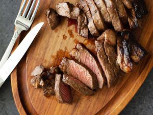 Overhead view of sliced steak cooked in the oven and silverware on a wooden cutting board.