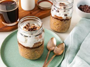 Jar of tiramisu overnight oats topped with Greek yogurt, mini chocolate chips, and cocoa powder on a small plate with two spoons, and in the surroundings, a bowl of mini chocolate chips, another jar of overnight oats, and a wooden board with a glass of coffee, a glass of milk, and the jar lid