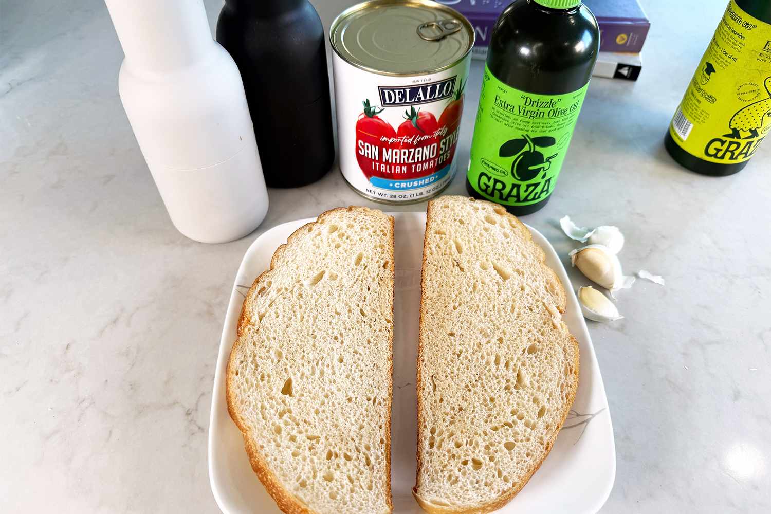 Ingredients arranged on a table including sliced bread on a plate tomato sauce can garlic cloves and bottles of oil and vinegar
