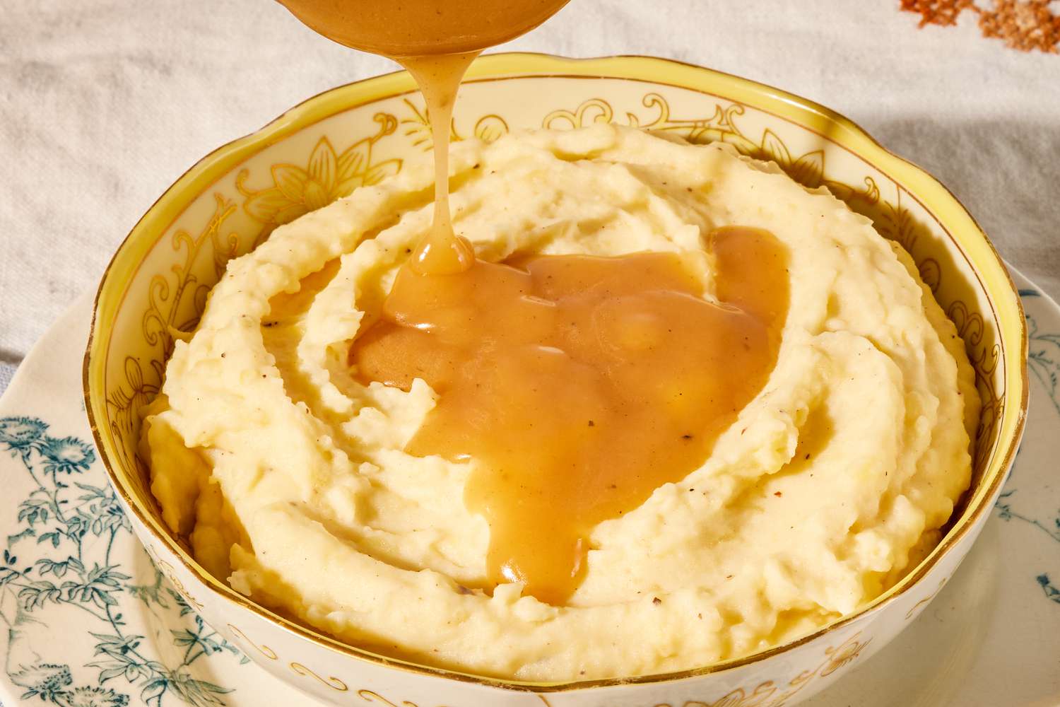 Angled view of a decorative bowl of mashed potatoes topped with gravy and sitting on a plate that is on a white and brown embroidered table cloth