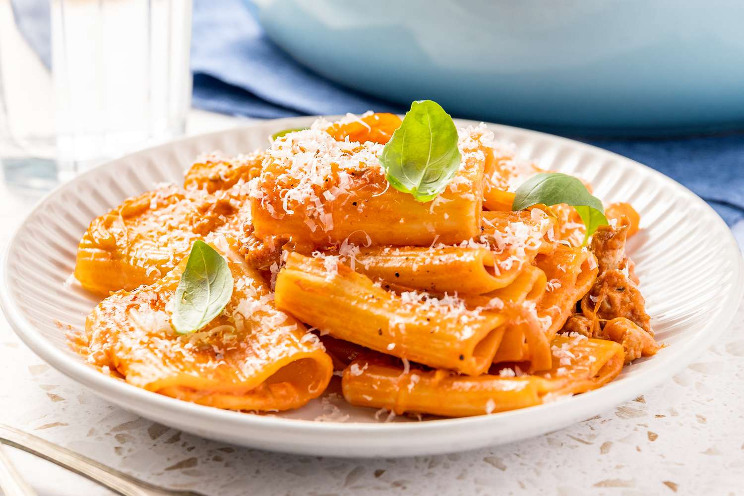 Plate of Paccheri With Quick Sausage Ragu Topped With Basil Leaves, and in the Surroundings, a Glass of Water and a Pot of More Pasta