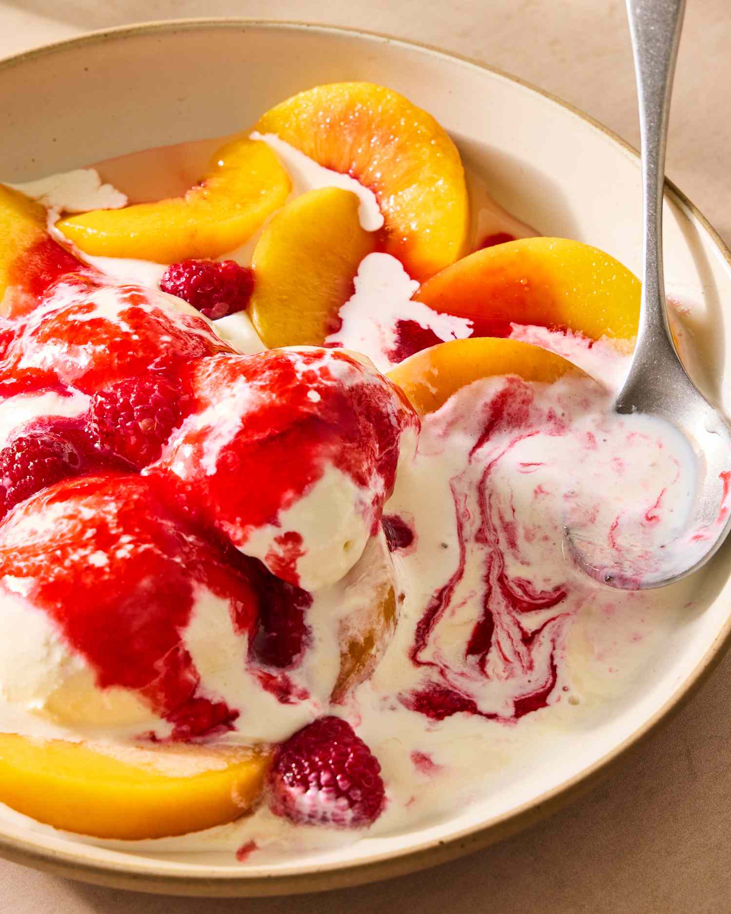 A bowl with peach slices, raspberries, ice cream, and raspberry sauce, with a spoon resting in it