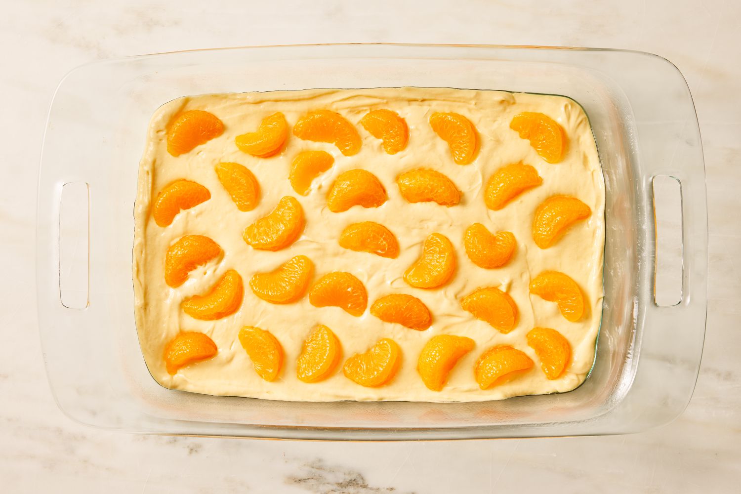 Overhead view of a glass baking dish of mandarin orange slices on top of cake batter for Pig Pickin' Cake recipe