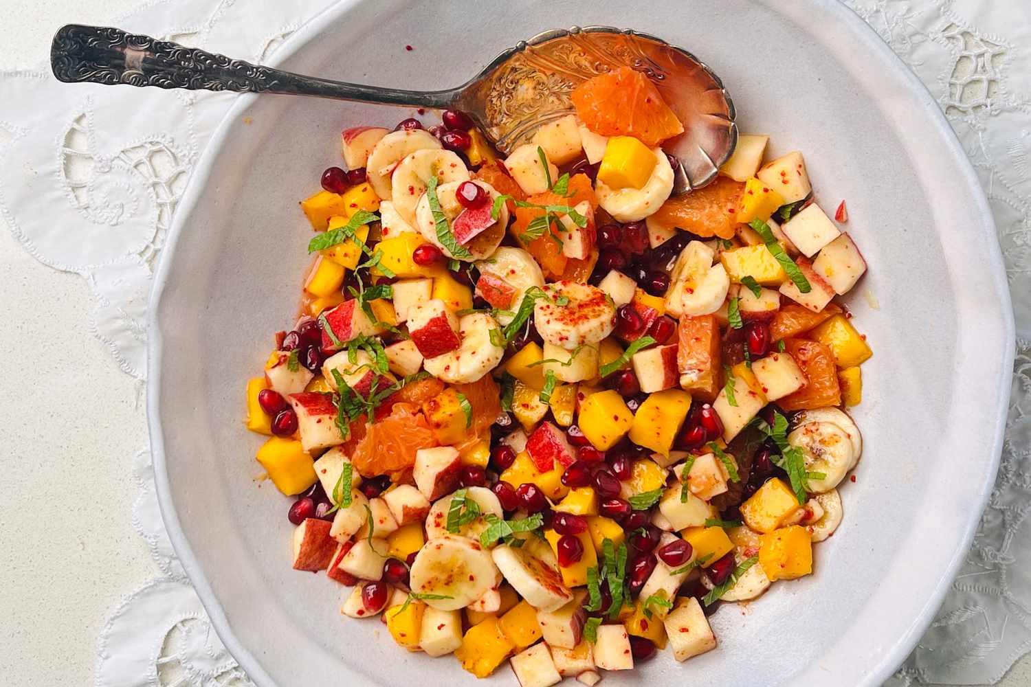 Fruit salad in a white bowl, showing banana slices, chopped apple, pomegranate arils, diced mango, and orange segments.