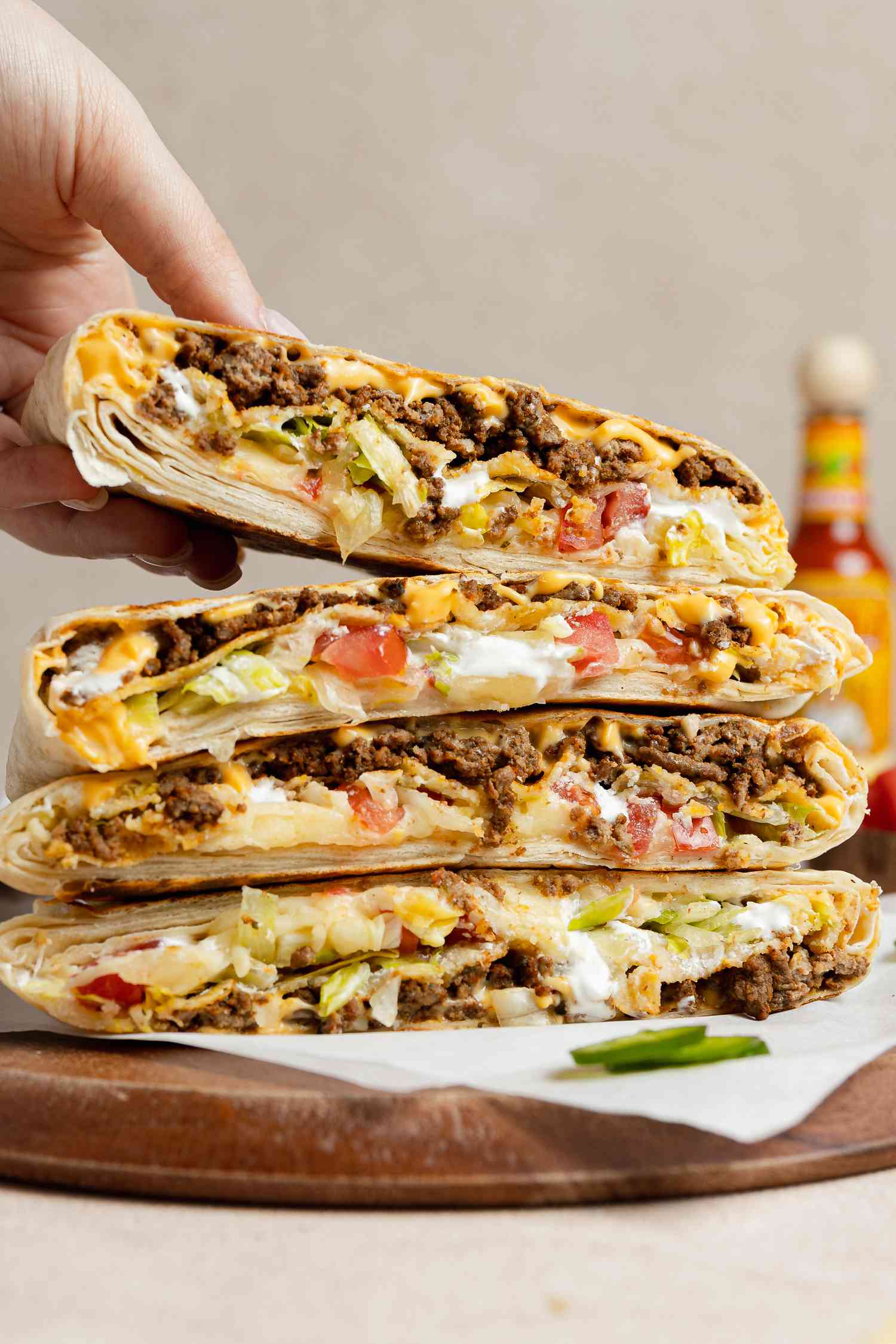 Hand Removing a Wrap from a Stack of Copycat Crunchwrap Supreme Wraps Cut in Half on Parchment Paper Over a Wooden Board