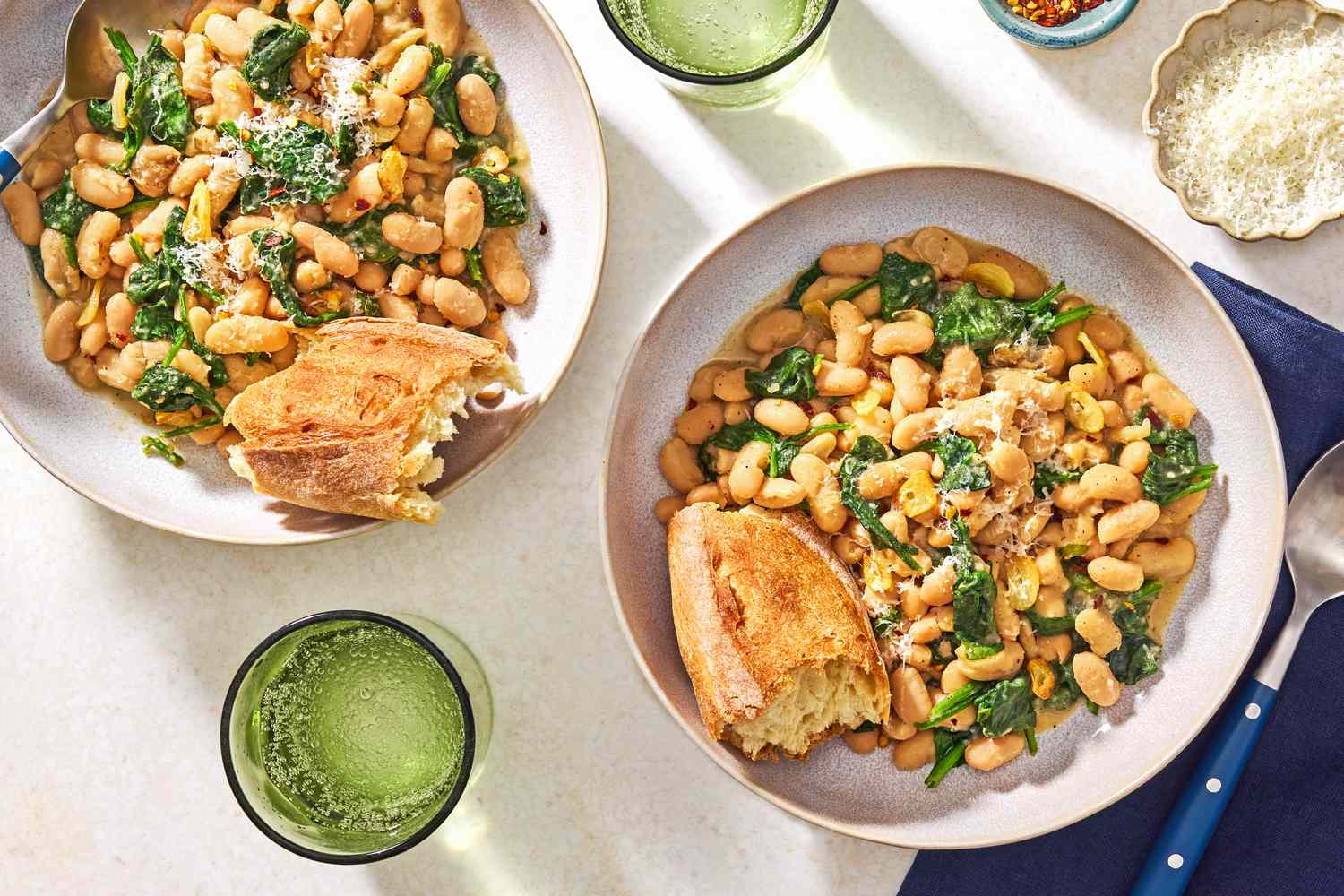 Overhead view of a bowl of beans and greens with a chunk of bread all next to a spoon and resting on a blue cloth napkin