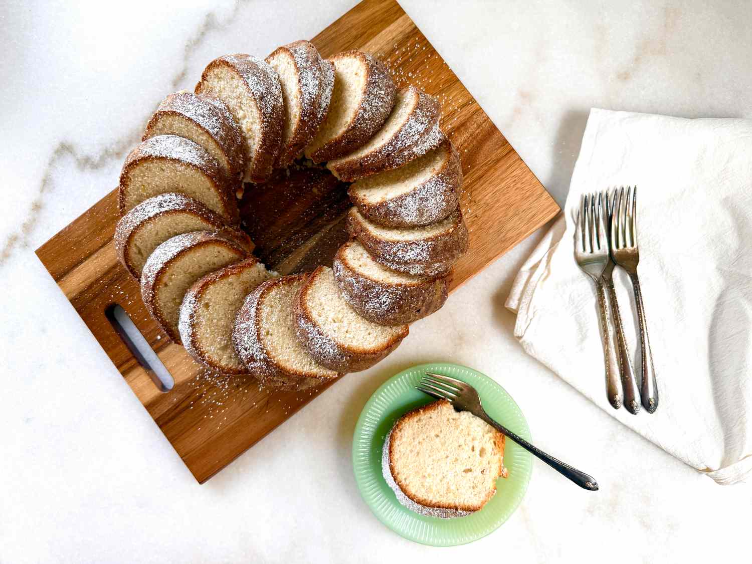 A ring-shaped almond cake sliced and displayed on a wooden tray, a plate with a slice and a fork beside a stack of utensils on a napkin